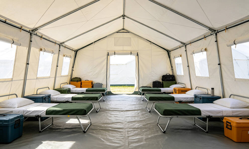 Emergency frame tent interior with organized rows of folding cots and clear walking aisles for shelter occupancy