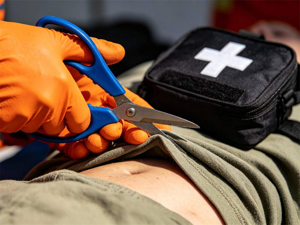 Close up action shot of orange gloved hand using blue trauma shears to cut clothing fabric with black medical pouch white cross nearby first aid training bright lighting