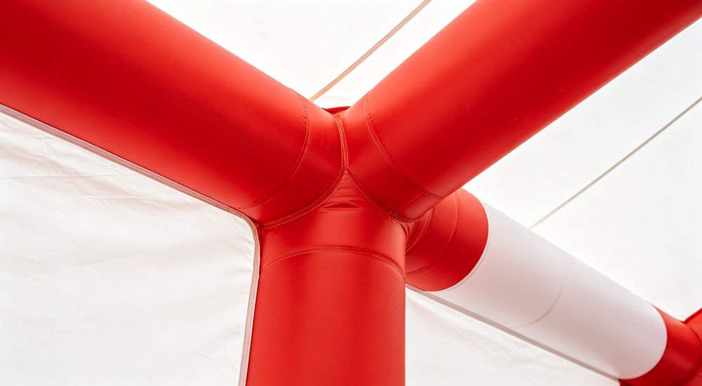 Side by side comparison of red and white inflatable tent with air beams vs green frame tent with metal poles, both on green grass, blue sky, professional product comparison for B2B buyers