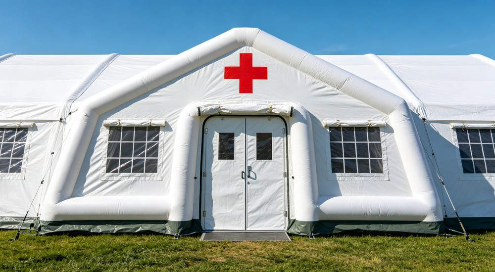 large white hospital medical inflatable tent with double entry doors, side windows, edge guy ropes, bold red cross sign on front gable and reinforced inflatable frame