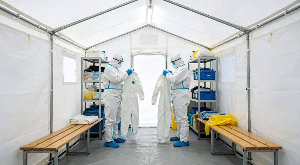Interior view of clean zone in decontamination tent with benches and storage shelves and workers removing protective suits in bright white interior