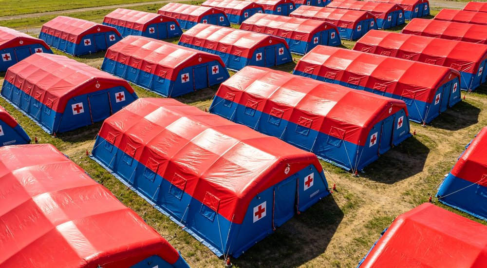 Aerial view of field hospital camp with rows of red and white inflatable medical tents, green grass, blue sky, medical workers
