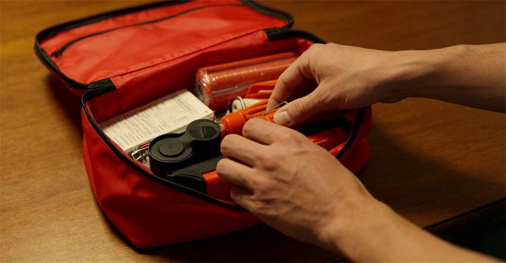 Close-up of hands packing small fire emergency kit supplies into waterproof red nylon bag with organized compartments on wooden table surface