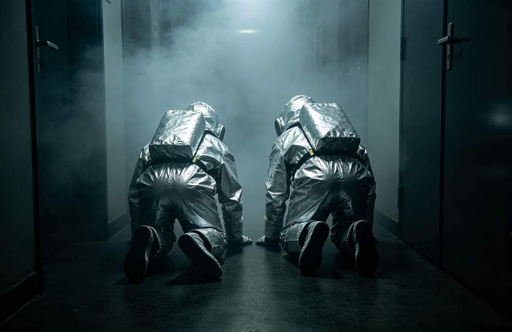 Two people wearing silver EN403 smoke masks crawling on floor to escape burning building with thick smoke during home fire evacuation drill today