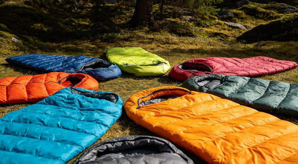Wide angle view of colorful sleeping bag collection in various colors, product variety display, outdoor natural setting with trees