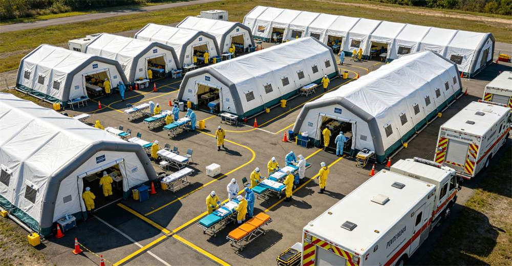Large scale mass casualty decontamination facility with multiple connected inflatable tents arranged in organized one way flow layout and medical triage area with emergency response vehicles present at the site