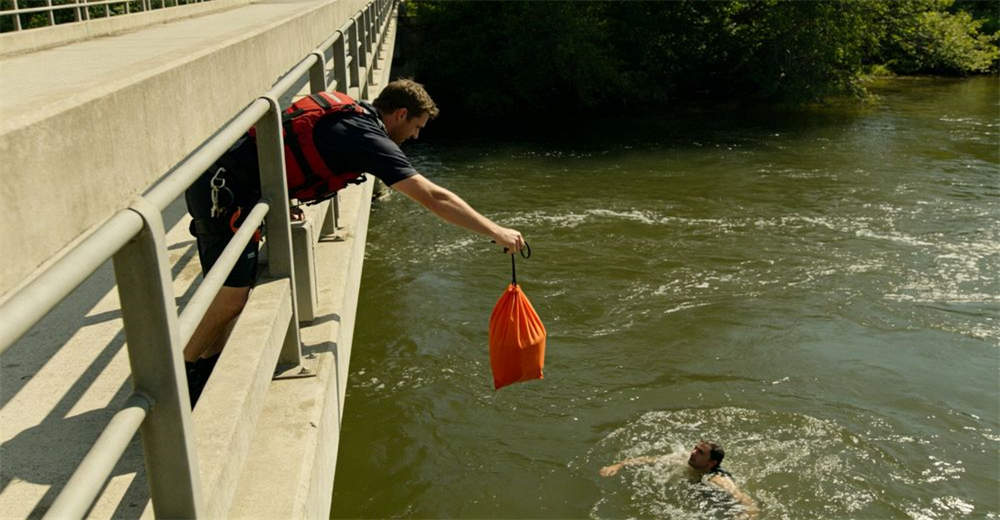 Action shot of dock worker rescue team throwing orange throw bag from concrete bridge railing down to water below showing real-world deployment conditions and team coordination