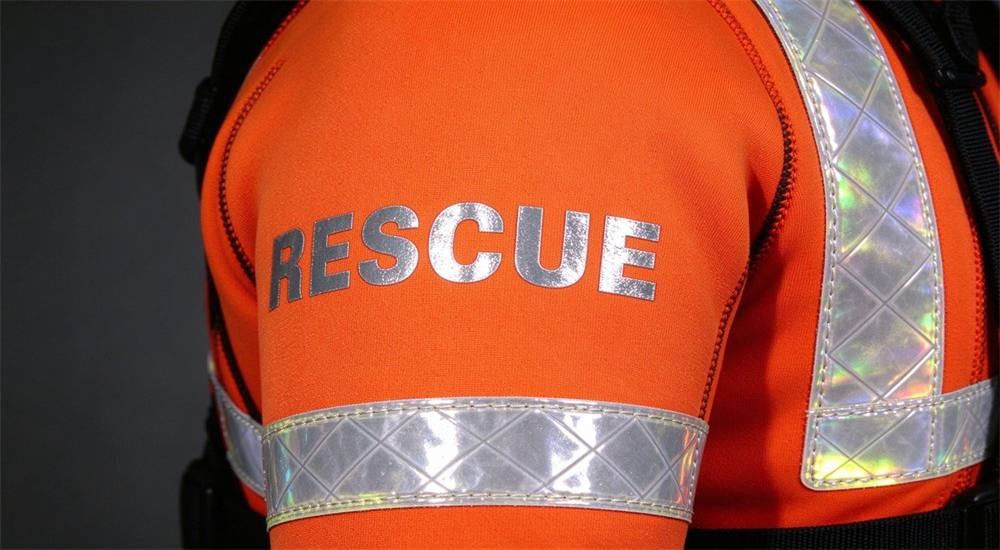 Close-up product detail of reflective RESCUE lettering printed on upper arm of orange neoprene wetsuit with high visibility reflective tape for low light water rescue identification Close-up product detail of reflective RESCUE lettering printed on upper arm of orange neoprene wetsuit with high visibility reflective tape for low light water rescue identification