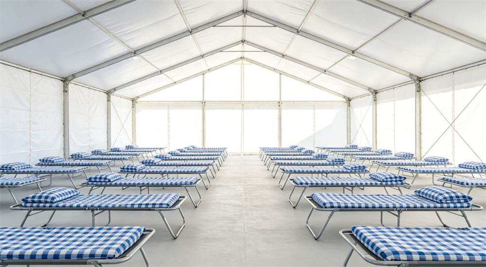 Front view of bright open aid tent interior with rows of camp beds dressed in blue and white check relief bedding, clean humanitarian emergency relief bedding ready for displaced families arriving after a disaster.