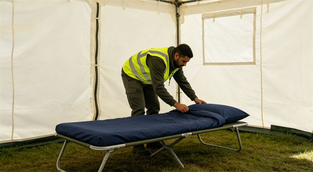 Overhead view of relief worker holding folded navy blue 140 GSM emergency relief bedding sets, standard weight humanitarian linen ready for distribution to displaced families in camps, in a warehouse supply chain for humanitarian aid operations.