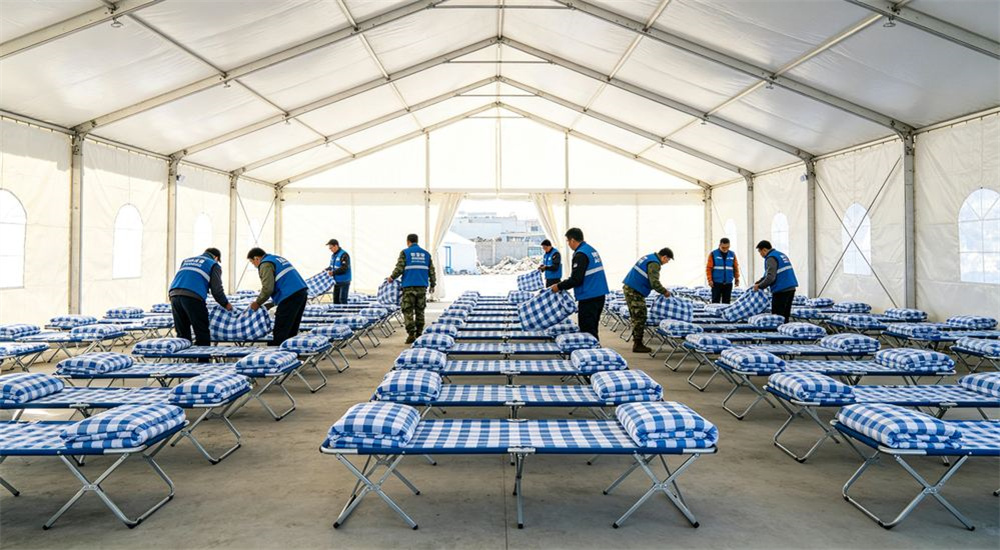 Wide-angle view of aid tent after an earthquake fitted with camp beds dressed in blue and white check relief bedding, rapid humanitarian emergency relief bedding for earthquake response operations, in a temporary camp set up immediately after a seismic disaster.