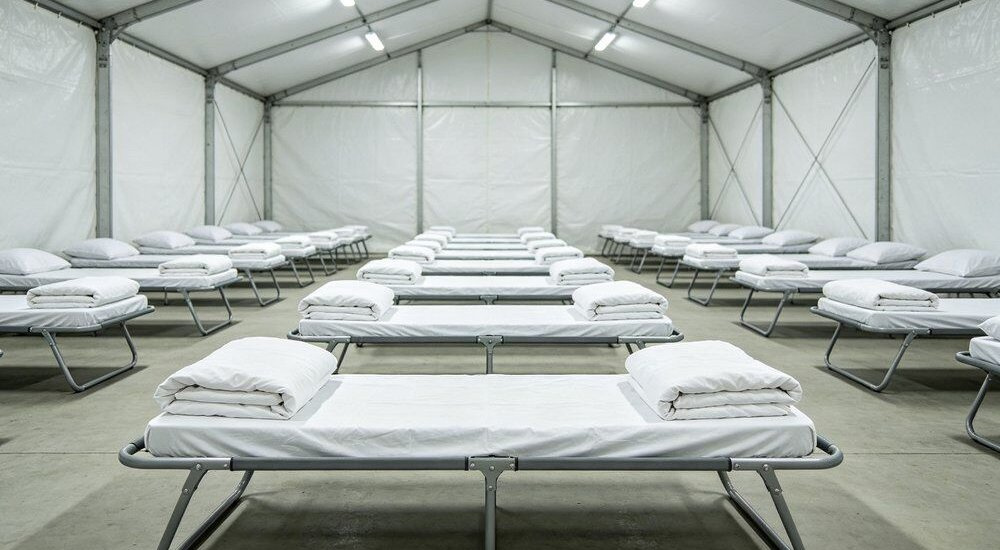 Aerial view of emergency relief shelter scene with neatly folded institutional bulk bedding sets arranged on metal cots, light blue cotton-poly sheet duvet cover and pillowcase sets ready for wholesale distribution across temporary disaster response camps