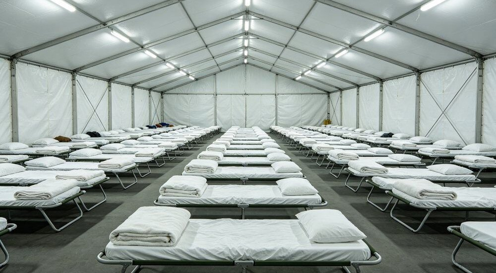 Wide-angle view of large emergency shelter scene with rows of blue institutional bulk bedding on cots in a humanitarian camp, wholesale relief linen ready for mass displaced person distribution in a tent hall