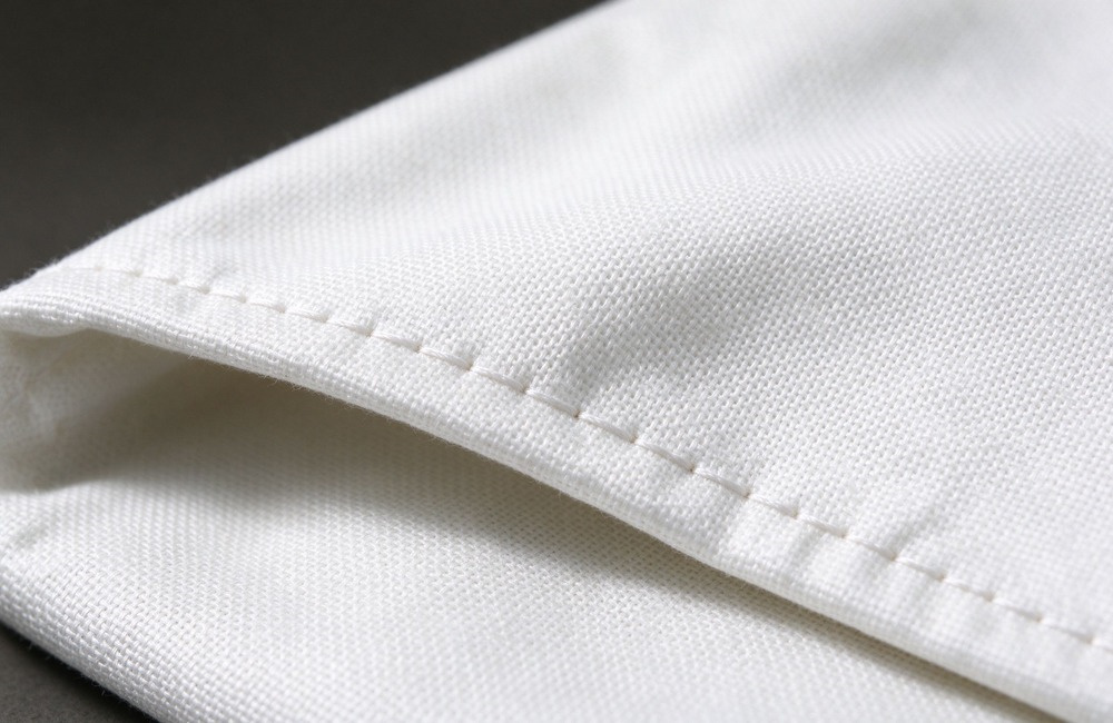 Extreme macro of the folded edge of a white fiberglass fire blanket showing tight white stitched binding, woven fiber texture and a black corner pull tab, soft even studio light