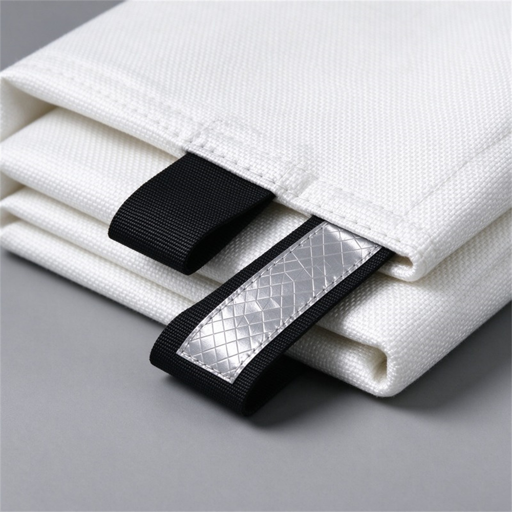 Square close-up of a black woven pull tab strap sewn onto the folded corner of a white fire blanket, white stitched binding visible, hand gripping the tab, light gray background Square close-up of a black woven pull tab strap sewn onto the folded corner of a white fire blanket, white stitched binding visible, hand gripping the tab, light gray background