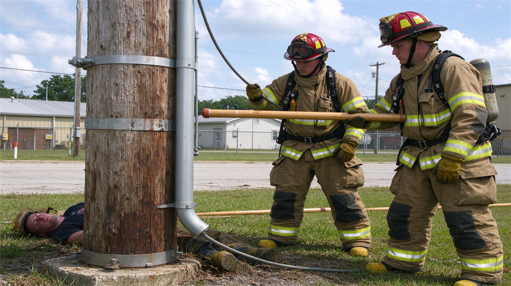 Fire training yard scene with two Caucasian firefighters in turnout gear practicing safe approach to a downed simulated power line pole, one holding a long wooden insulating pole, daytime training ground, different male faces