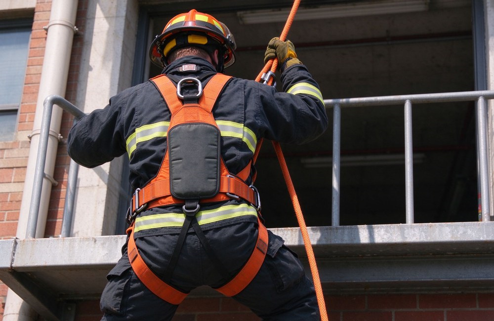 Medium rear view shot of a Western firefighter in dark turnout gear and orange full body safety harness standing on a building ledge gripping an orange kernmantle fire escape rope, back D-ring and EVA padding clearly visible, ready to descend Medium rear view shot of a Western firefighter in dark turnout gear and orange full body safety harness standing on a building ledge gripping an orange kernmantle fire escape rope, back D-ring and EVA padding clearly visible, ready to descend