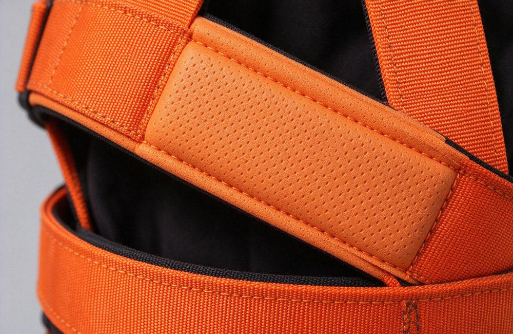 Close-up detail shot of the shoulder strap and leg loop of an orange and black full body safety harness showing the open-cell EVA foam padding stitched under the polyester webbing, steel adjustable buckle visible Close-up detail shot of the shoulder strap and leg loop of an orange and black full body safety harness showing the open-cell EVA foam padding stitched under the polyester webbing, steel adjustable buckle visible