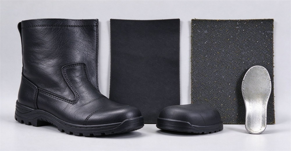Pair of black tall PVC rubber safety boots with silver steel toe caps and yellow side indicator tag, orange hard hat placed beside them on light gray studio background