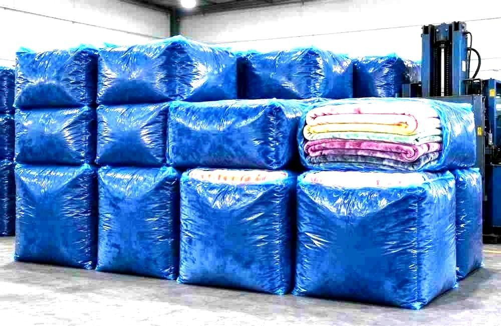 Product view of compressed flannel blanket bales wrapped in transparent film with visible dense stacked layers, warehouse lighting, organised rows